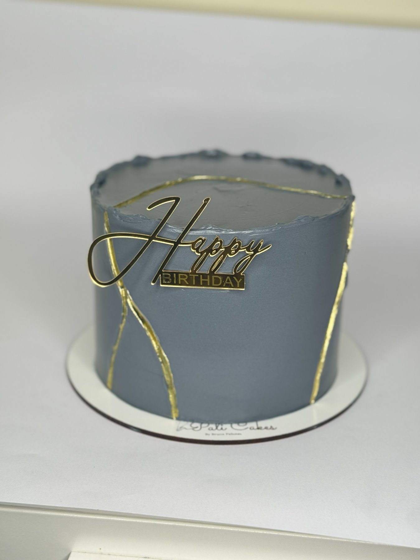 Bolo cinzento com linhas douradas em efeito kintsugi e topo dourado de aniversário produzido pela Pali Cakes